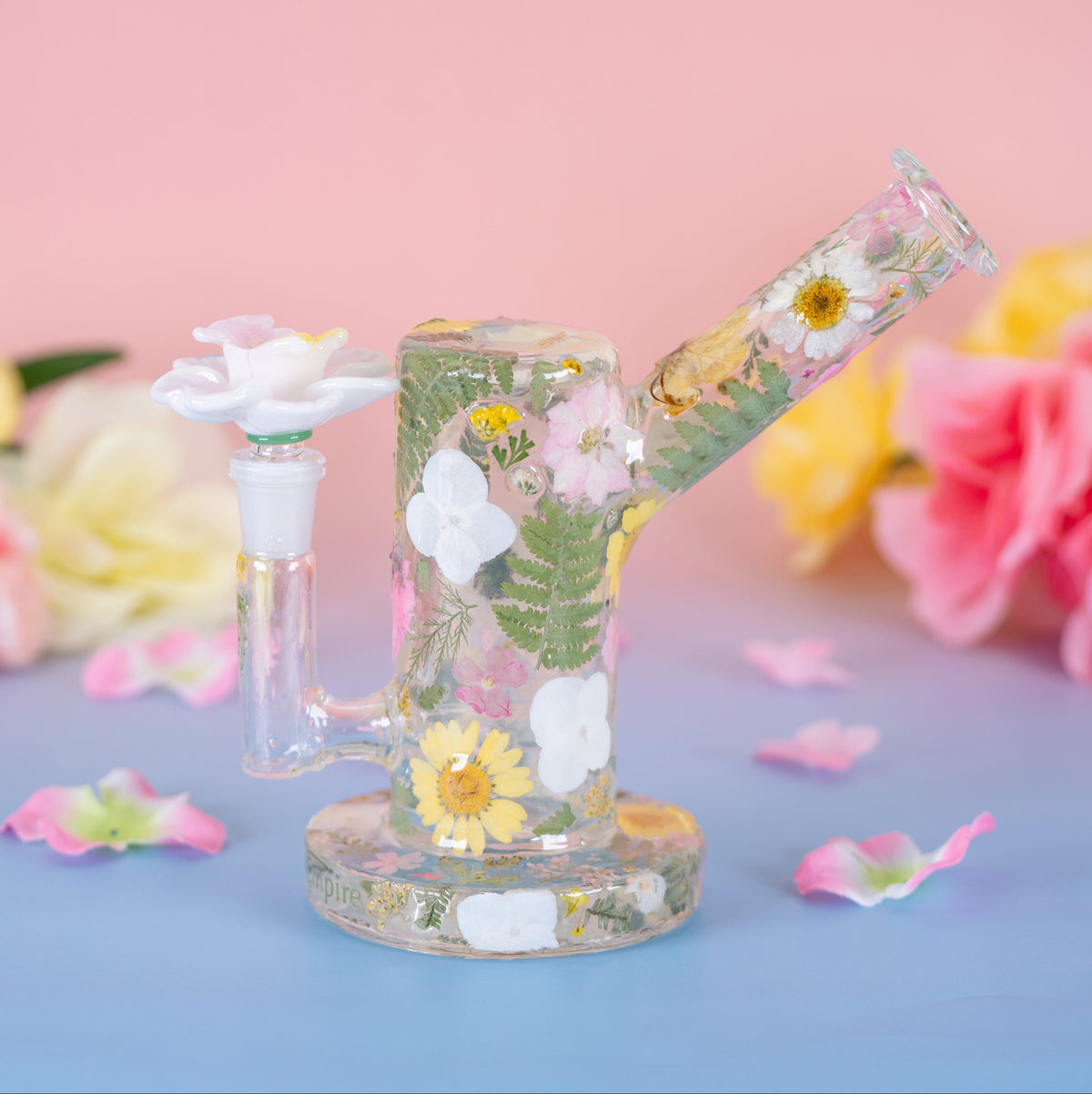 A clear glass water pipe with embedded dried flowers and green leaves, displayed on a blue surface with a pink background and artificial flowers.