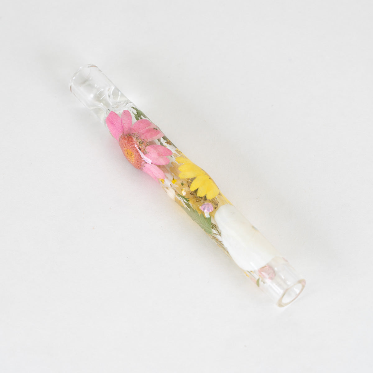 A clear glass smoking pipe containing pressed flowers, including pink and yellow blossoms, set against a plain white background.