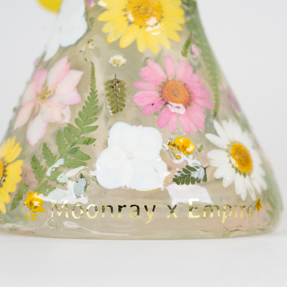 A close-up of a decorative glass vessel with a conical shape, decorated with pressed flowers and green leaves in yellow, pink, white, and green, with gold foil text reading 'Moonray x Empire' near the base.