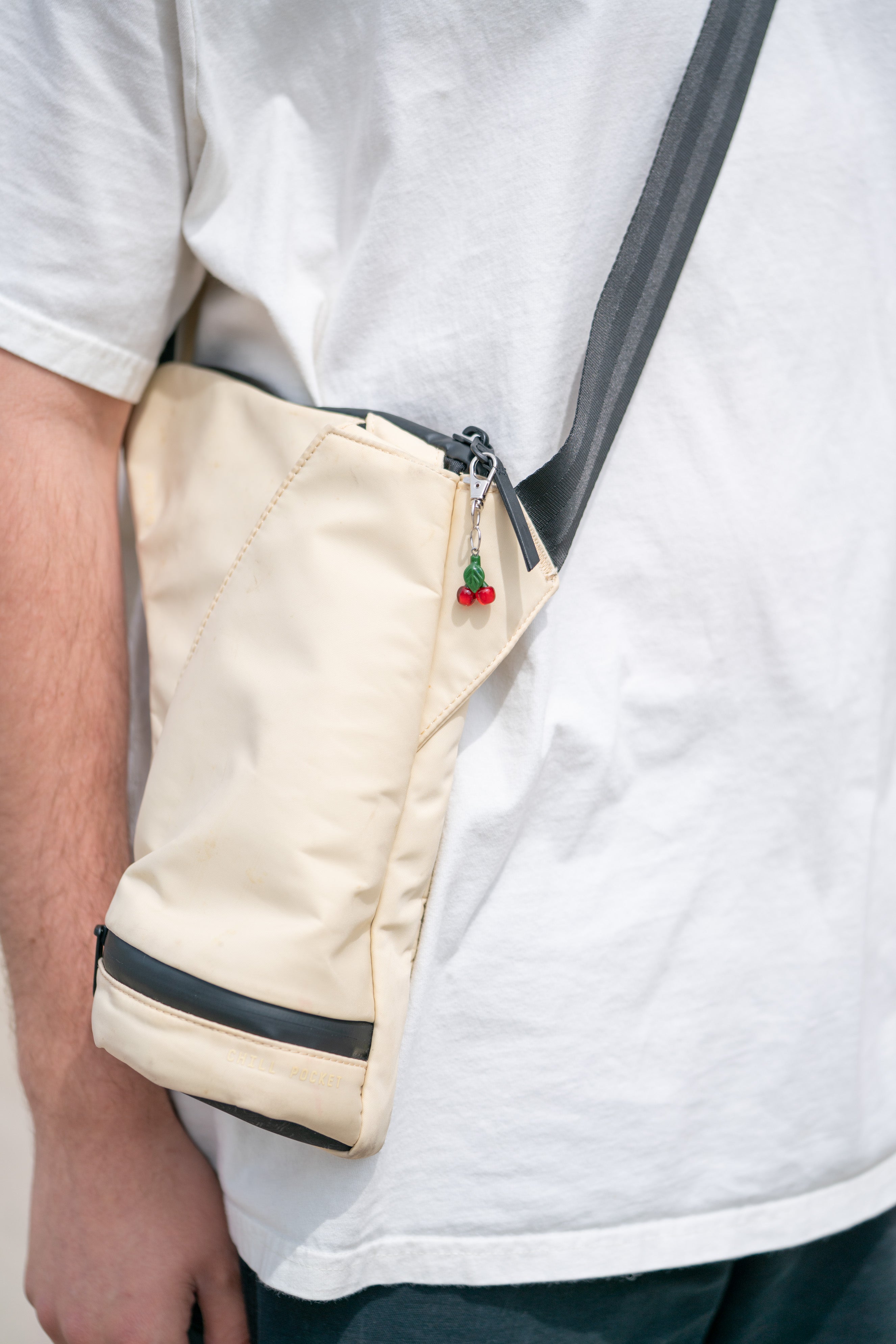 Beige crossbody bag with a small red and green ornament on a white shirt background