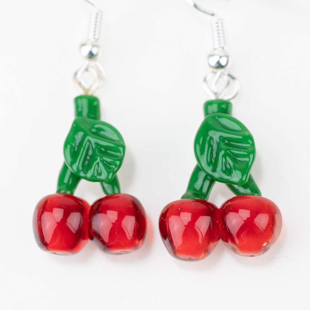 A close of a set of cherry dangle earrings. Each earring has two cherries and a stem with leaves. The cherries are a deep red and the stem is green.