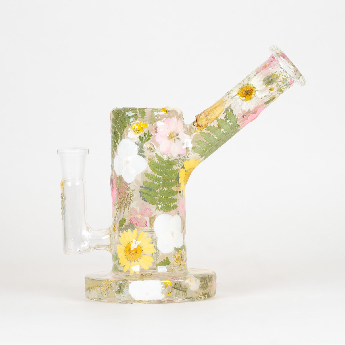 A clear glass water pipe with a rounded base and angled mouthpiece, decorated with pressed flowers and green leaves in pink, yellow, white, and green tones, set against a plain white background.
