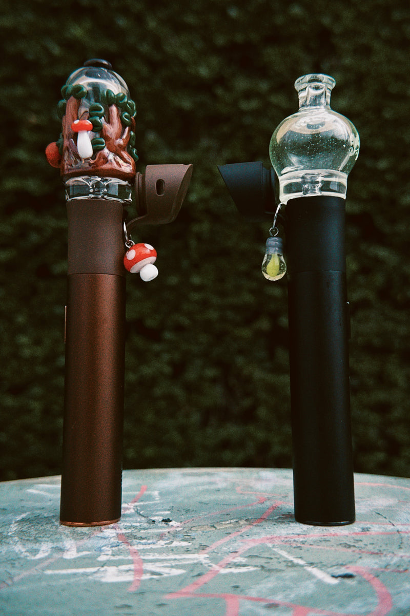 Two smoking accessories, one with a decorative top and one plain, on a textured surface with a dark background.