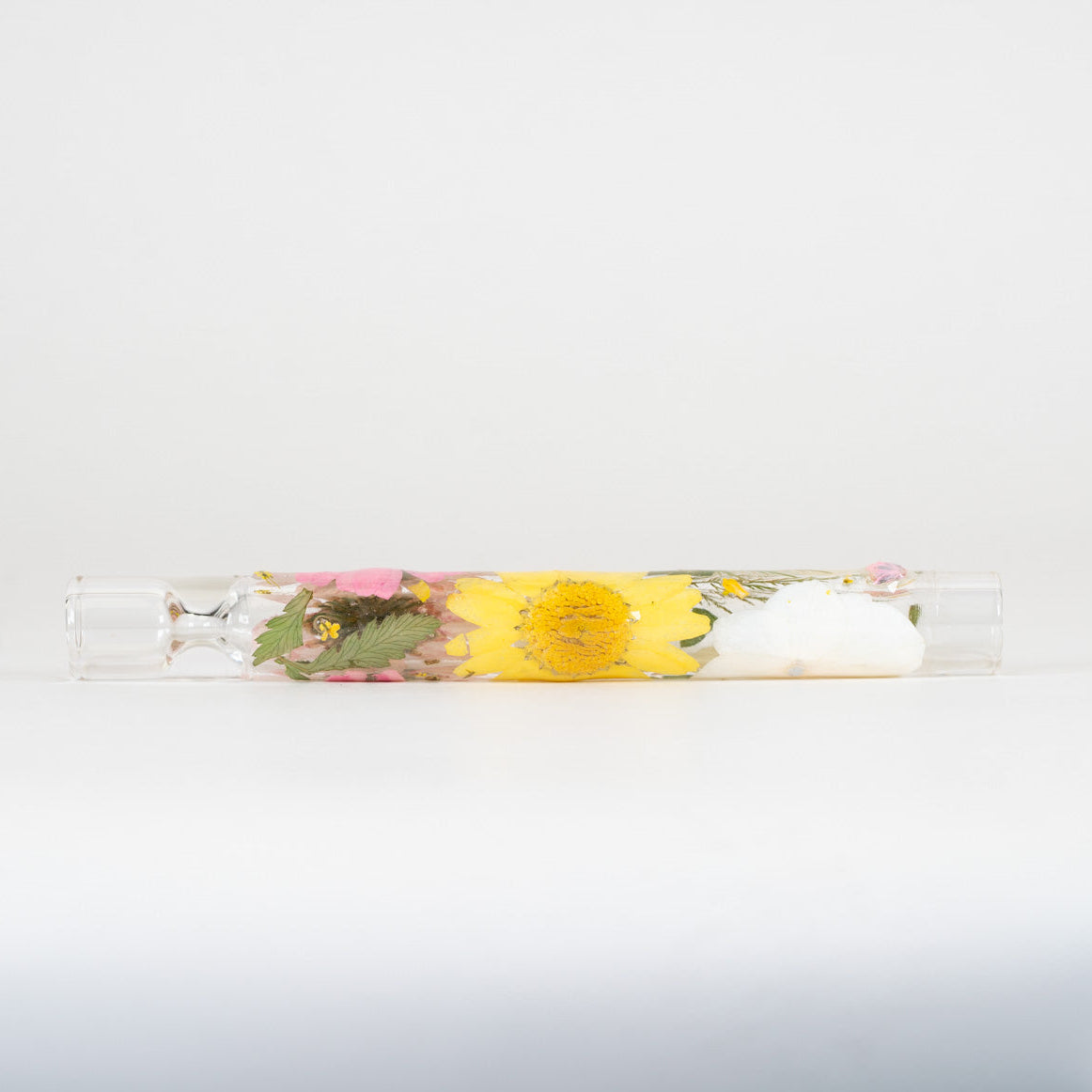 A clear cylindrical glass tube containing a yellow flower, pink blossoms, green leaves, and a white wax seal, photographed horizontally against a plain white background.