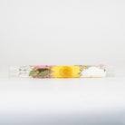 A clear cylindrical glass tube containing a yellow flower, pink blossoms, green leaves, and a white wax seal, photographed horizontally against a plain white background.
