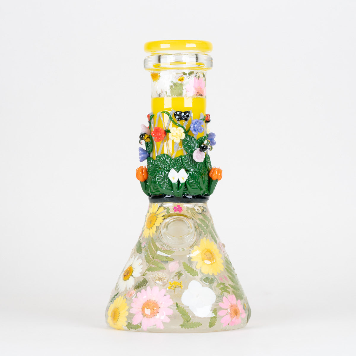 A clear glass beaker with a yellow lid, decorated with pressed flowers, green leaves, and yellow sunflowers on a white background.