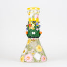 A clear glass beaker with a yellow lid, decorated with pressed flowers, green leaves, and yellow sunflowers on a white background.