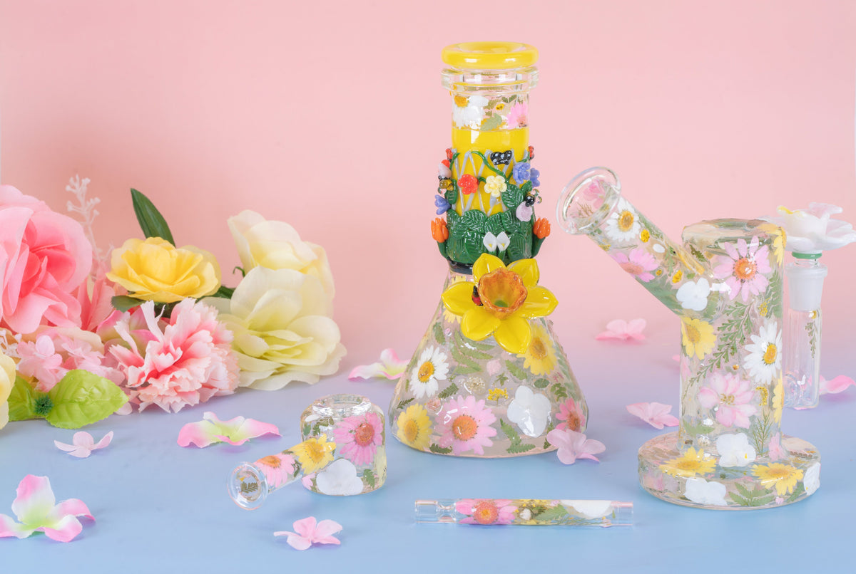 A series of floral glass smoking products that are covered in dried flowers encased in resin. The collection includes a baby beaker bong, a dry spoon pipe, a chillum, and a mini rig all with floral elements.