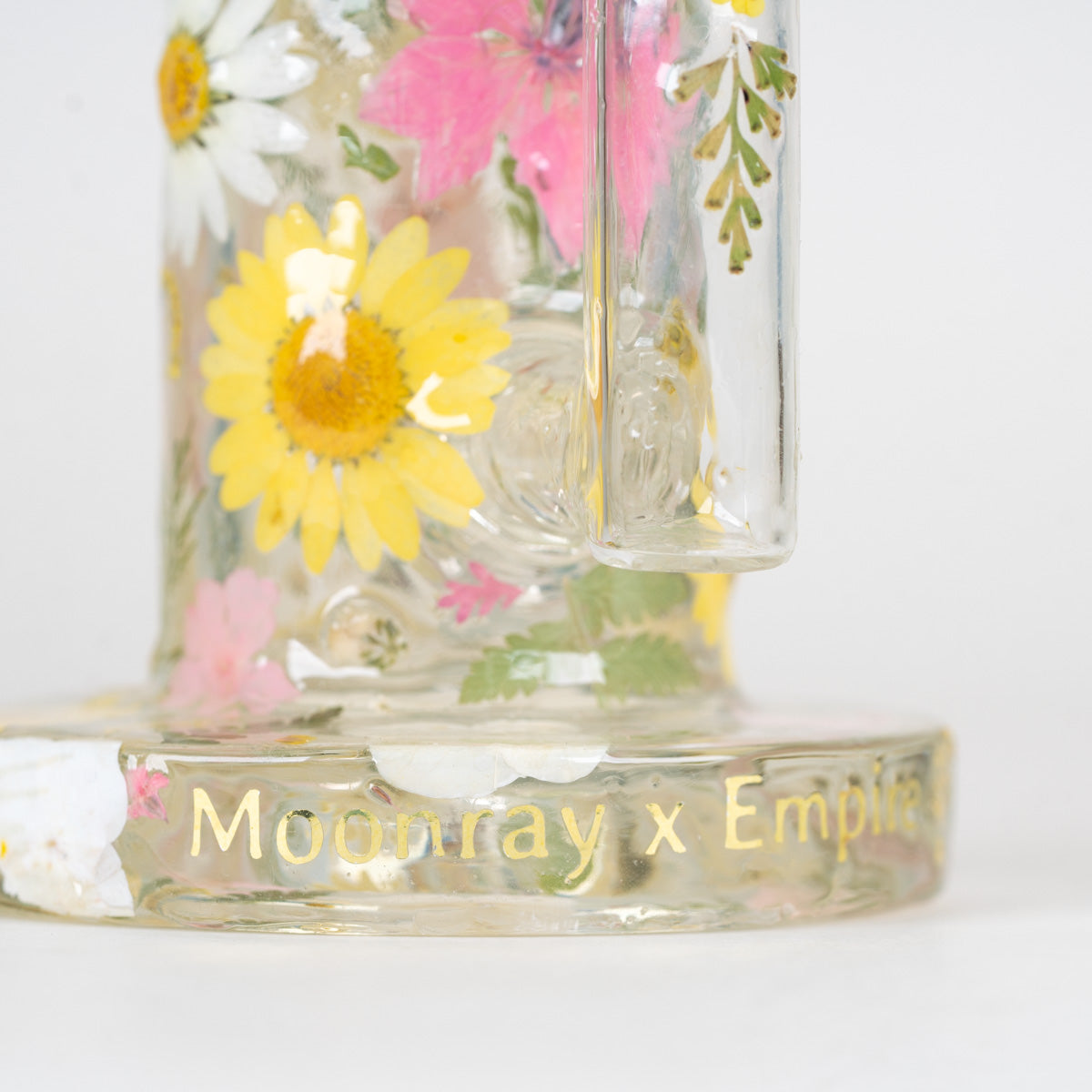 A close-up of a clear glass banger with pressed floral decals, including a prominent yellow daisy and pink flowers, and gold foil text reading 'Moonray x Empire' on the base, set against a plain white background.