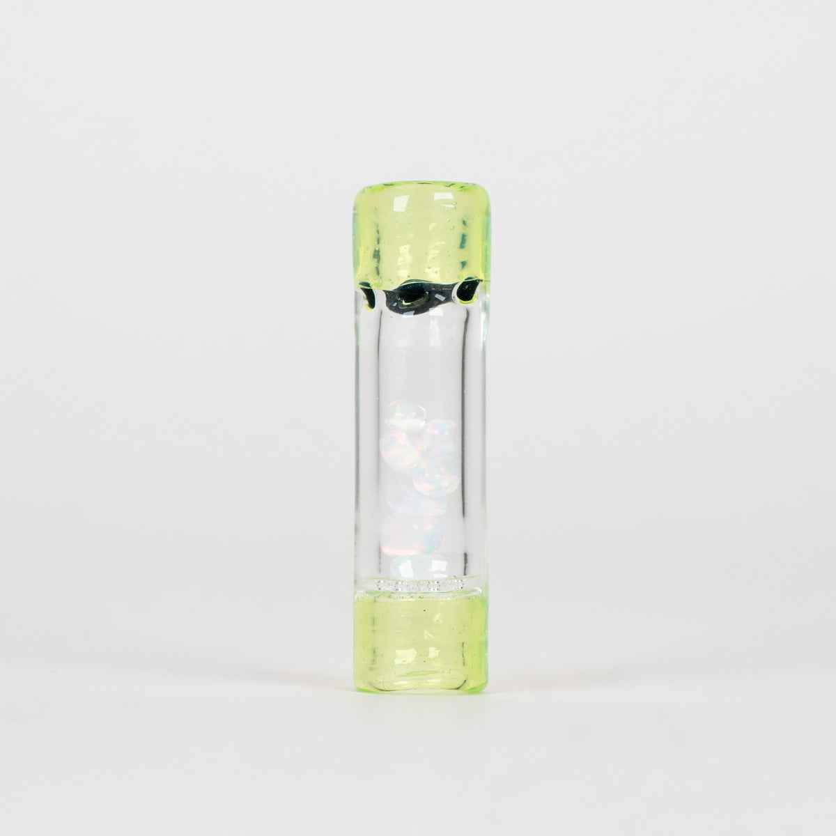 A clear cylindrical glass container with a translucent light green cap and base, featuring a black printed graphic and a white iridescent figure inside, set against a plain white background.