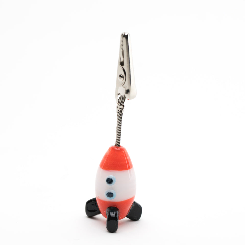A roach clip with a rocket shape base, holding a joint. The Spaceship is red and white with black wings.