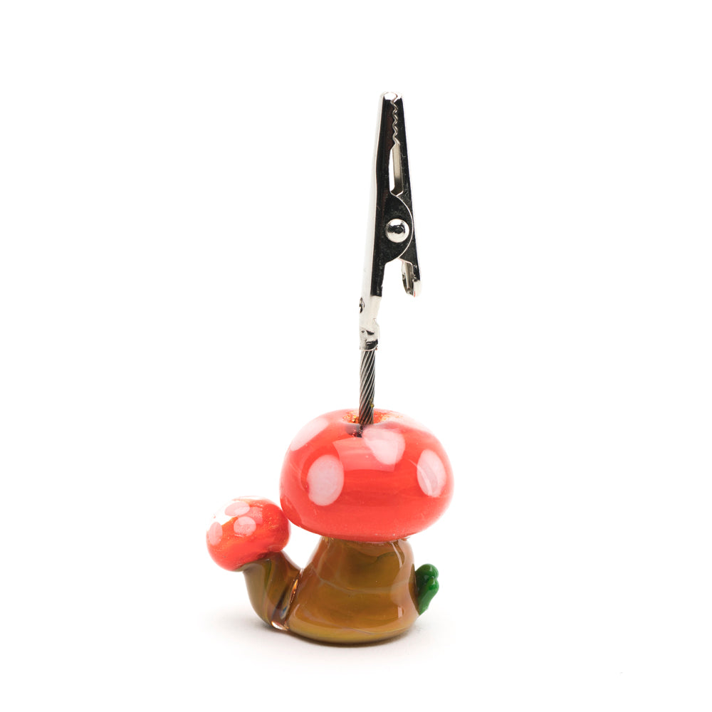 A roach clip shaped like a mushroom used to hold joint. the mushroom is red with white dots and has a brown base. The roach clip portion is metal.